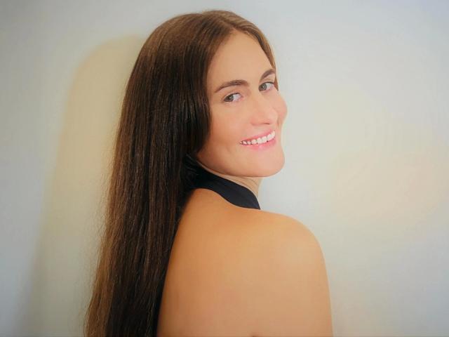 ReifeCecilla-hot has long brown hair, smiling with pink lips, looking over her shoulder, showing her bare back and black neck strap, with fair skin and brown eyes.