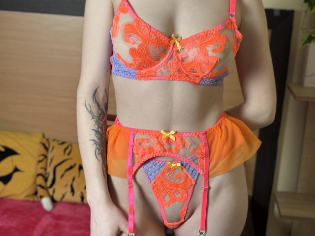 HappyTina wearing bright orange and purple lace lingerie with pink straps and yellow bows, showing her tits and pussy. She has a tattoo on her left arm.