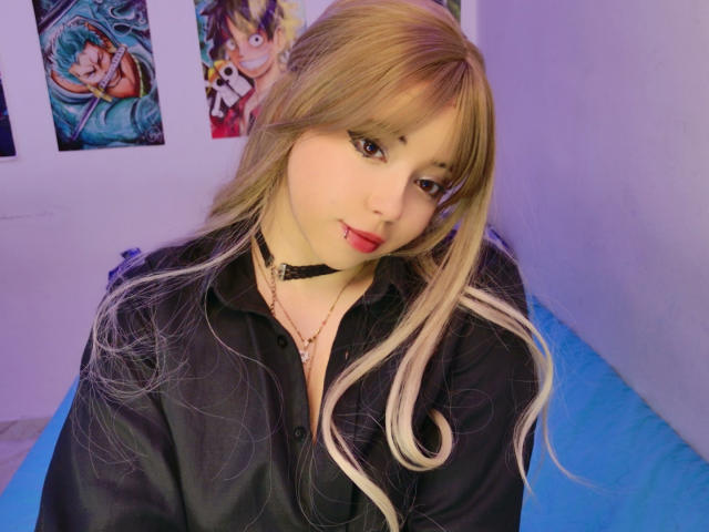Arieyaara, con cabello rubio y blanco, un collar negro, una camisa negra y lápiz labial rojo, sentada en un sofá azul, mirando a la cámara con una ligera sonrisa..
