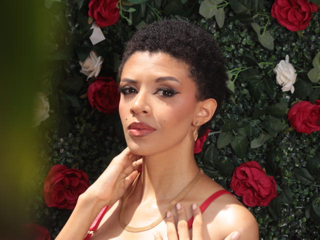 SamantaRoa tem cabelo preto encaracolado e curto, pele morena e batom vermelho, vestindo um vestido vermelho com brincos dourados, com as mãos tocando o pescoço, cercada por rosas vermelhas e brancas..