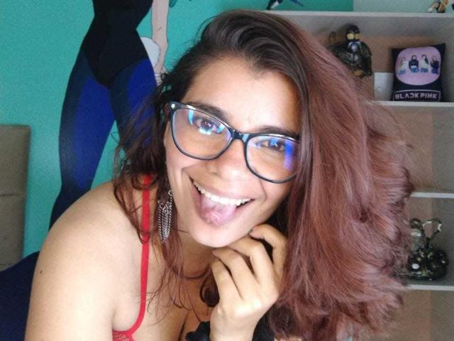 BulmaHot with red hair, glasses, and tongue out, smiling, showing her tits, wearing a red top, and big dangly earrings, in a selfie.