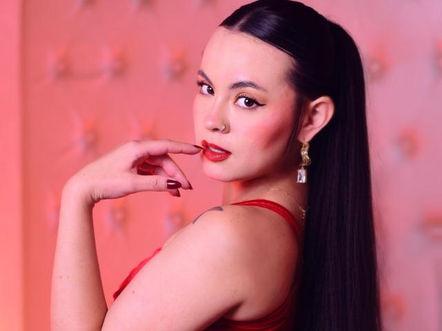 Ariana Rossy, con lunghi capelli neri, rossetto rosso e orecchini rossi, morde il dito, mostrando il suo reggiseno rosso e un piccolo tatuaggio sulla spalla. Il suo seno è visibile..
