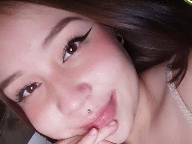Moonat has a cute, pinkish face with brown eyes, winged eyeliner, a nose piercing, a lip piercing, and is biting her finger with a subtle smile.