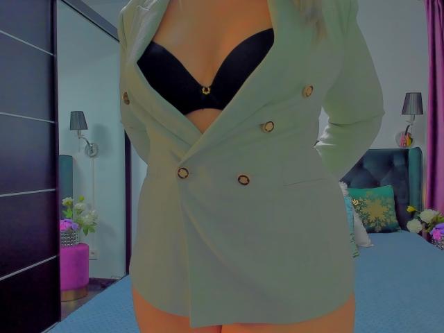 HotKrystall usando um blazer verde de corte duplo, sutiã preto com logo dourado, mãos nos bolsos, mostrando os seios, em pé em um quarto com cama azul e vaso rosa..