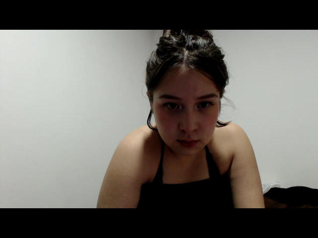 Moonat with dark hair in a bun, wearing a black halter top, has a nose ring and is looking down with a serious expression, sitting against a white wall.