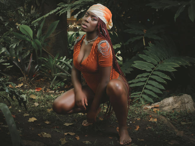 AvaBelle, squatting, wearing an orange dress with deep cleavage, and a white-orange headwrap, in a forest, with dark brown skin, braids, and bare feet.