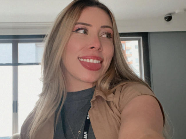 JennyLawry, avec ses cheveux blonds, son rouge à lèvres rouge et son sourire, la langue légèrement sortie, portant une chemise beige par-dessus un haut gris, regardant sur le côté dans un autoportrait..