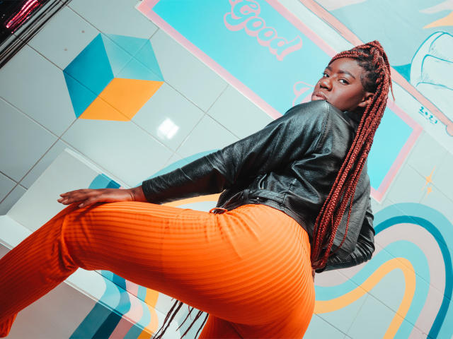 AvaBelle met donkere huid, rode vlechten, een zwarte leren jas en oranje geribde broek, kijkt om, met haar hand op haar knie, haar bil iets omhoog, in een helder gekleurde, moderne omgeving..
