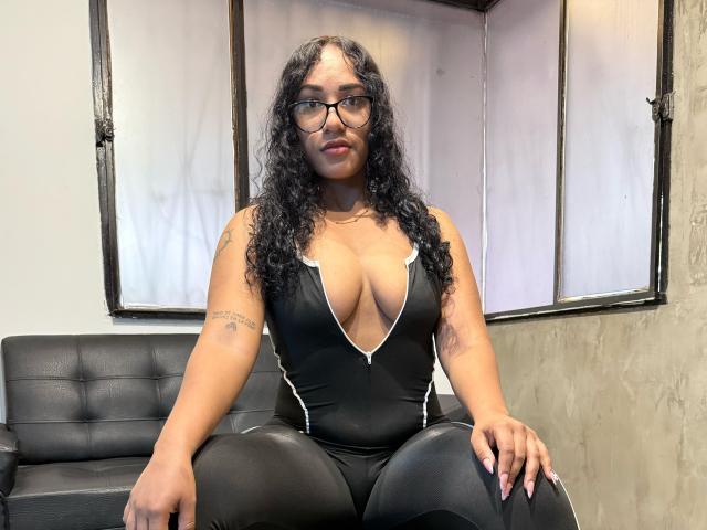 ThiainaRose, con cabello negro rizado, gafas y un body negro de cuello en V pronunciado, que muestra sus grandes senos y su brazo tatuado, sentada en un sofá de cuero negro..
