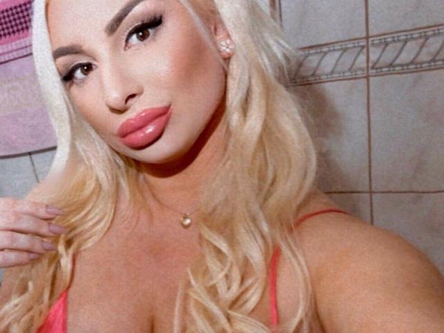 LuxuriaBlond has blonde hair, pink glossy lips, and is wearing a pink spaghetti strap top, showing her tits. She's in a bathroom with tiled walls.
