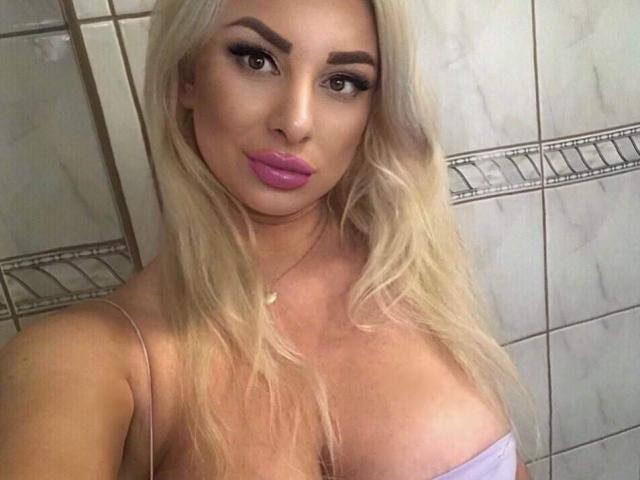 LuxuriaBlond with blonde hair, pink lipstick, and large tits in a white strap top, staring into the camera with a seductive look.