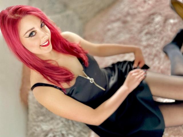 DominaLuna with bright pink hair, smiling, wearing a black dress with a front zipper, sitting on a pink fuzzy surface, showing her tits and holding a phone.
