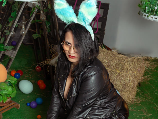 DonnaJones with black hair, wearing a leather jacket, blue fuzzy bunny ears, glasses, and red lipstick, sitting on green grass with colorful eggs and hay bales.