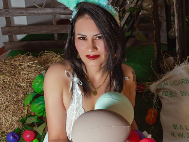 DonnaJones with dark hair, wearing white lace top, blue headband with cat ears, holding balloons, has red lipstick, gold necklace, and looks directly at camera with slight smile.