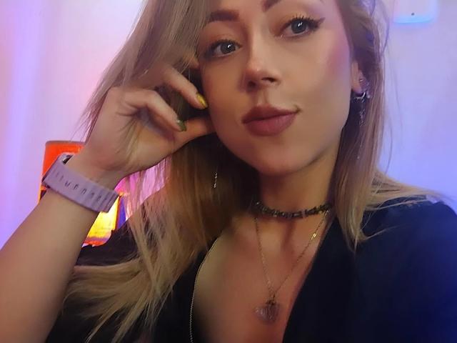 DouceKhrystine has blonde hair, wearing a black top, choker, and wristband, with yellow nails, and a heart necklace, looking at the camera with a slight smile.
