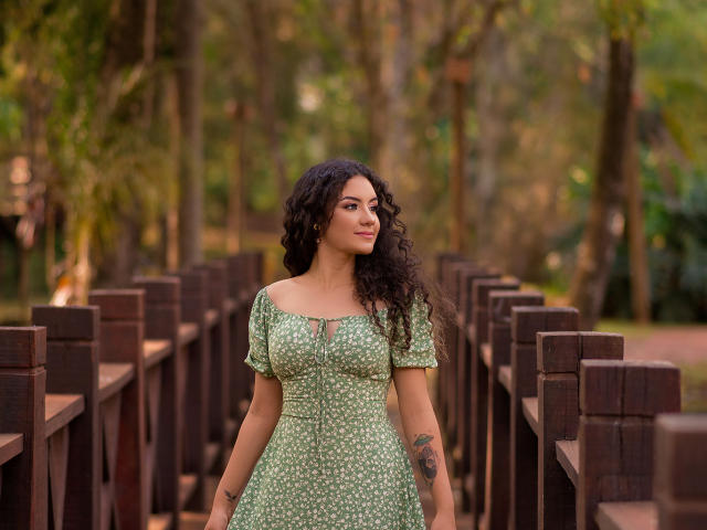 CaskiallX with curly black hair, green dress with white polka dots, small tits, tattoos on right arm, standing on wooden pathway, forest background, looking to the side, smiling.