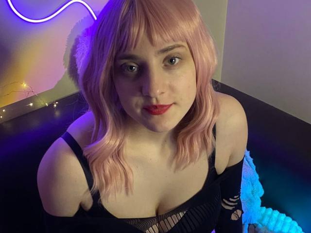 EmilieGold69 with pink hair, red lipstick, black mesh top, showing her tits, sitting in a dark room with neon lights and blue blanket. She's smiling at the camera.