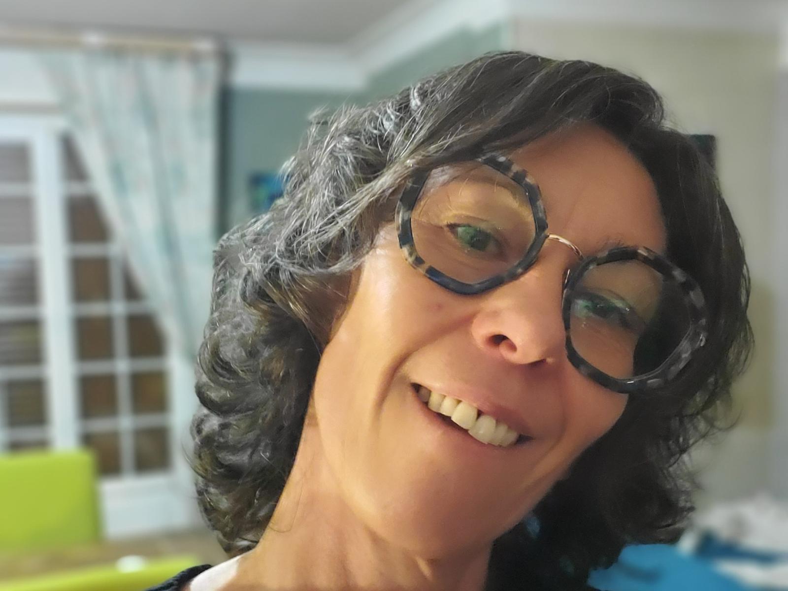 TileaStar, com cabelos pretos encaracolados, usando óculos redondos grandes, sorrindo mostrando os dentes, tem pele clara e está olhando ligeiramente para o lado em uma selfie casual..