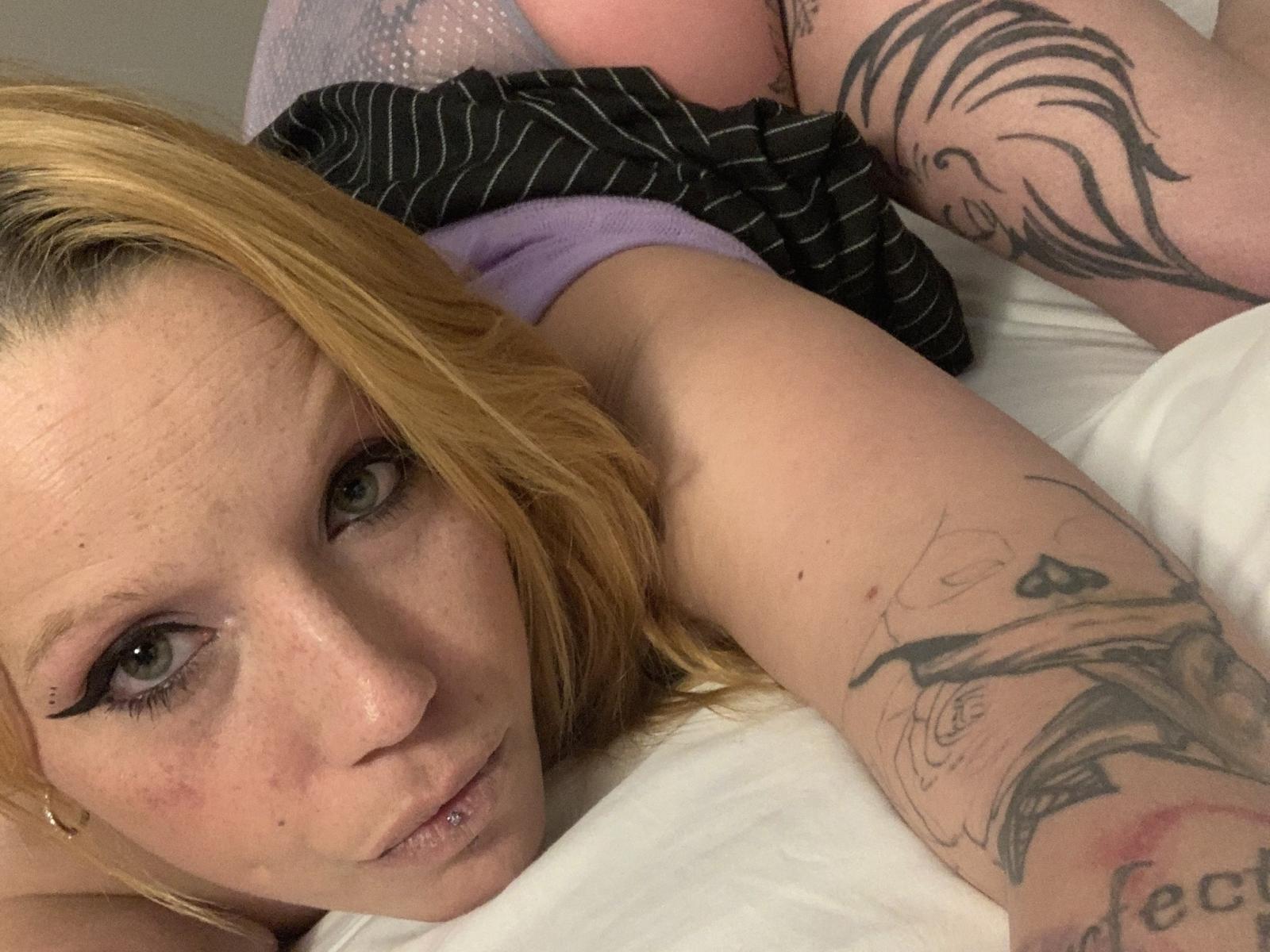 KandyCakee, blonde with tattoos, lying on bed, showing arm tattoos, pouty lips, green eyes, black eyeliner, and a slight nose piercing, wearing purple and black-striped clothes.