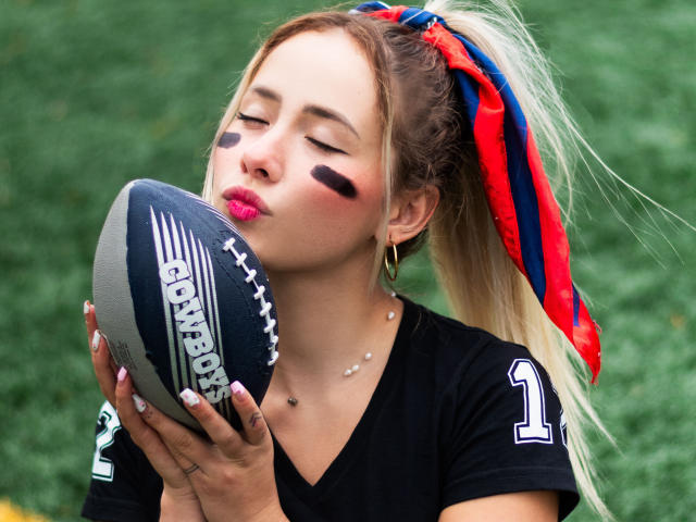 NatSorian sta baciando un pallone da calcio, con il viso dipinto di nero, indossa una maglia nera con una coda di cavallo rossa, blu e bianca, e una collana dorata. Le sue labbra sono dipinte di rosa..