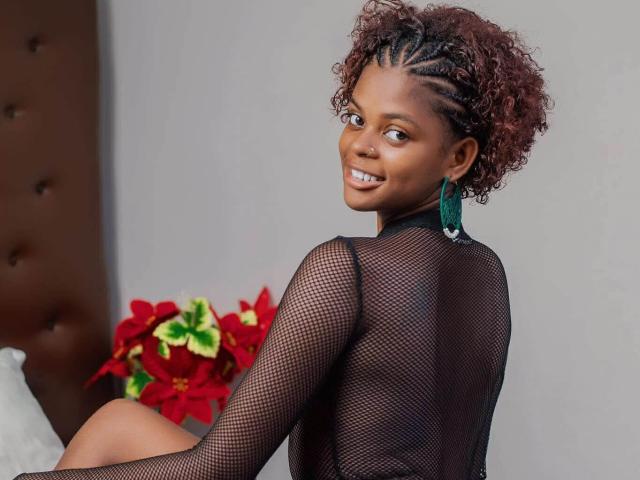 JessiccaHot with curly brown hair, braided sides, wearing a black fishnet top, green earrings, smiling, looking back, sitting on a bed with red and green poinsettias.