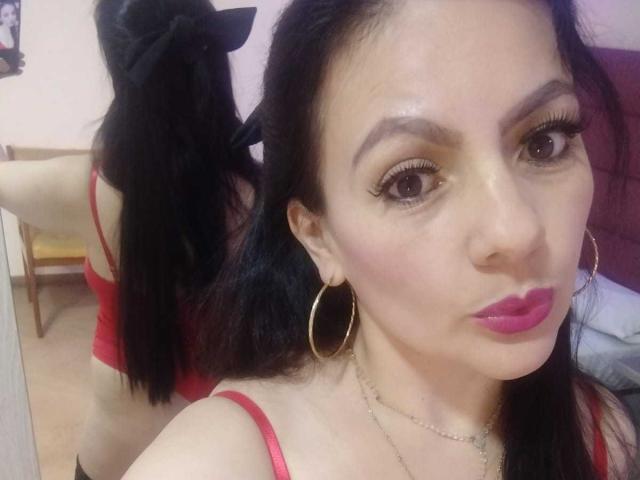 JakyKidman com cabelo preto, vestindo uma blusa vermelha, mostrando suas costas em um espelho, usando brincos de argola dourados, batom rosa e um laço preto, em uma selfie..