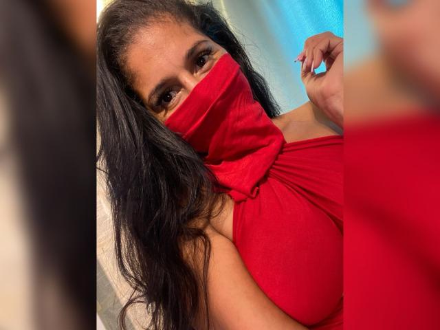 Kira Taleb, avec de longs cheveux noirs, portant un masque rouge et une robe rouge, qui couvre ses seins, sa main droite près de son visage, des ongles bleus, une expression concentrée, éclairage intérieur..