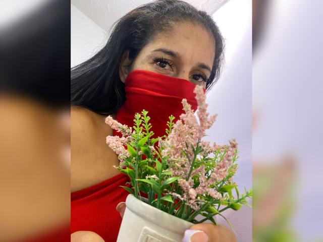 KiraTaleb with dark hair, wearing a red mask and red dress, holds a small pink flower pot, her tits barely visible, reflected in a mirror.