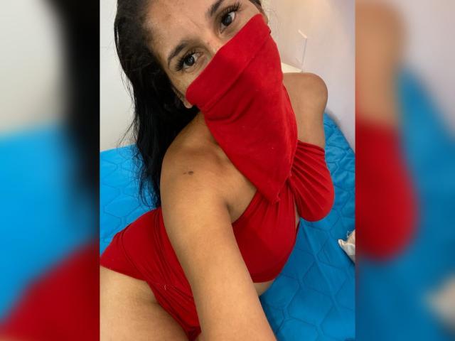 KiraTaleb with dark hair, red mask, and red top, showing her tits, wearing red shorts, on blue bed, reflection in mirror, brown eyes, medium skin, looking at camera.