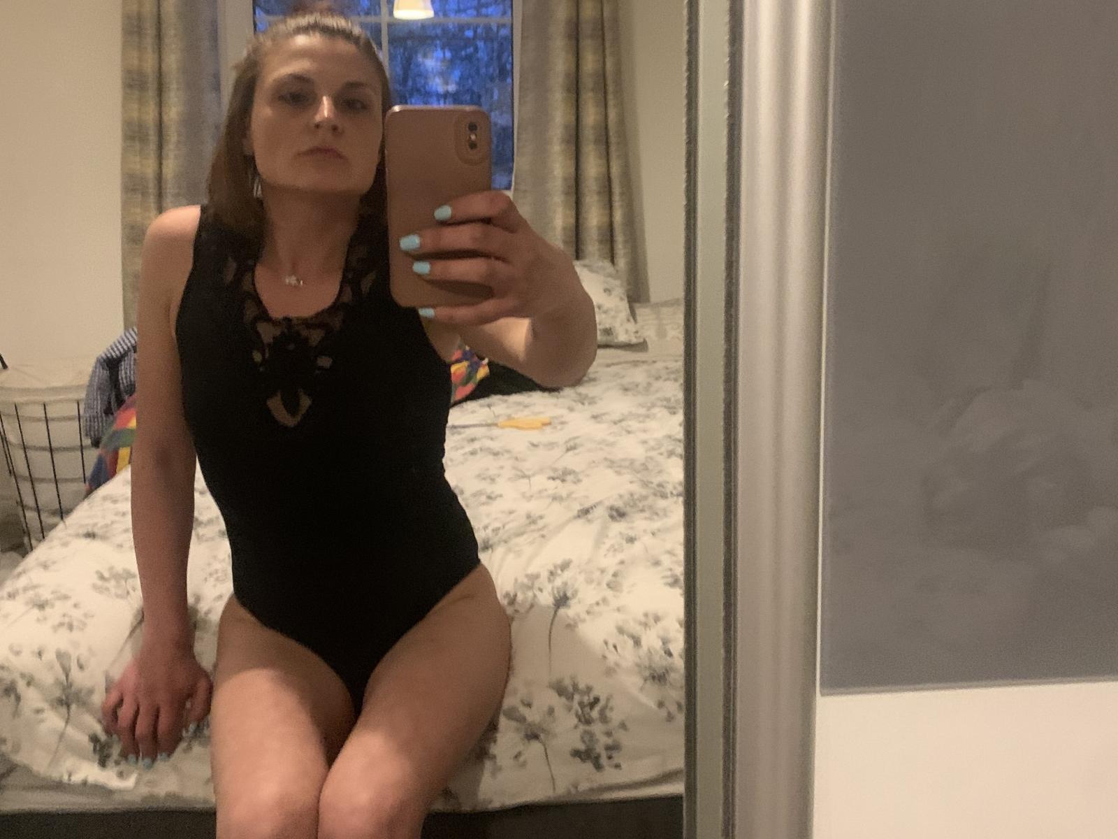 Tracey, mit braunen Haaren, trägt einen schwarzen, schnürbaren Body und sitzt auf einem Bett. Sie macht ein Selfie im Spiegel und zeigt dabei ihr Gesäß und ihre Beine, wobei ihre Nägel türkis sind..