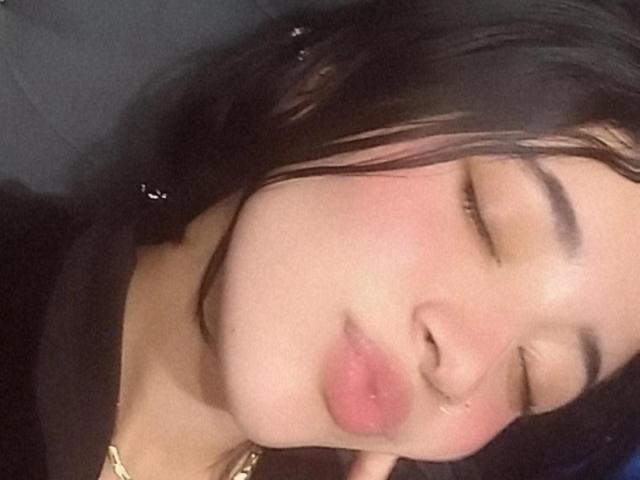 SophiiaFerrero's face, pouty pink lips, closed eyes, slight blush, black hair, soft skin, wearing a black top, gold necklace, relaxed expression, slight sheen on her face.