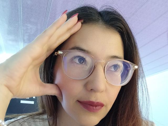 AntonnellaS with glasses, red nails, and pink lipstick, saluting with her right hand, showing her tits slightly, under a purple ceiling, looking at the camera.