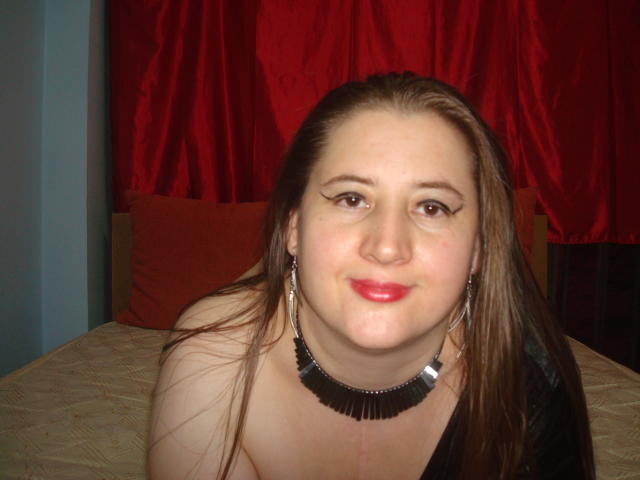 Superstar with long brown hair, red lipstick, and black fringed necklace, wearing earrings, sitting on bed, smiling, showing her tits, in a bedroom with red curtains.