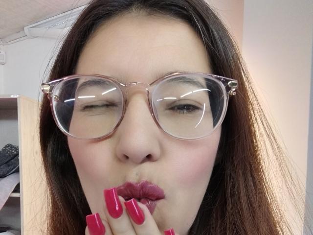 AntonnellaS with puffy pink lips, pink glossy nails, and clear glasses, sucking her fingers, brown hair, light skin, reflective glasses, slightly closed eyes, pink lipstick, in a selfie.