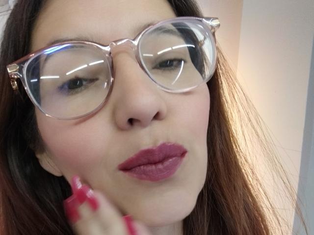 AntonnellaS with pink glasses, pouty purple lips, and pink nails, looking at camera with slightly parted lips. Brown hair, fair skin. Reflective glasses. Close-up.