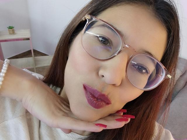 AntonnellaS with glasses, red lipstick, and pink nails, pouting, hand on cheek, wearing a pearl bracelet, looking at camera.