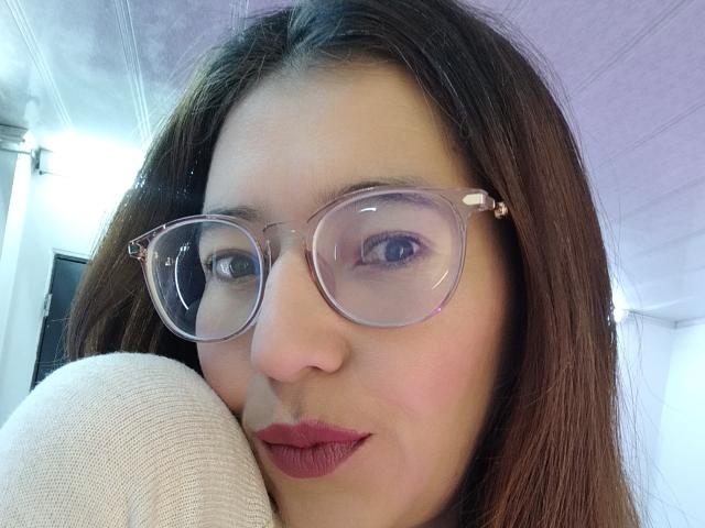 AntonnellaS with clear glasses, brown hair, and red lipstick, resting her head on a white sleeve, showing her cute face and slightly smiling. Her tits aren't visible.