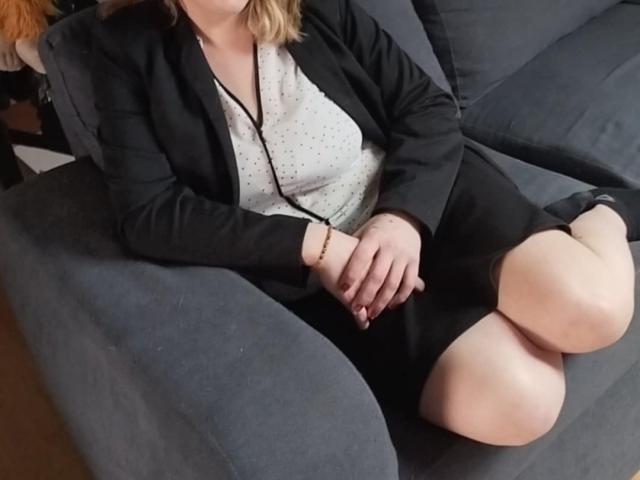 CoupleCadresPervertis with pale skin, blonde hair, black blazer, white polka dot blouse, black skirt, and bare legs, sitting on grey couch, hands resting on knee.