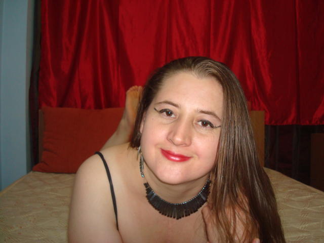 Superstar with long brown hair, red lipstick, black necklace, and bare shoulders, lying on a bed, smiling at the camera, showing her tits. Red curtains in the background.