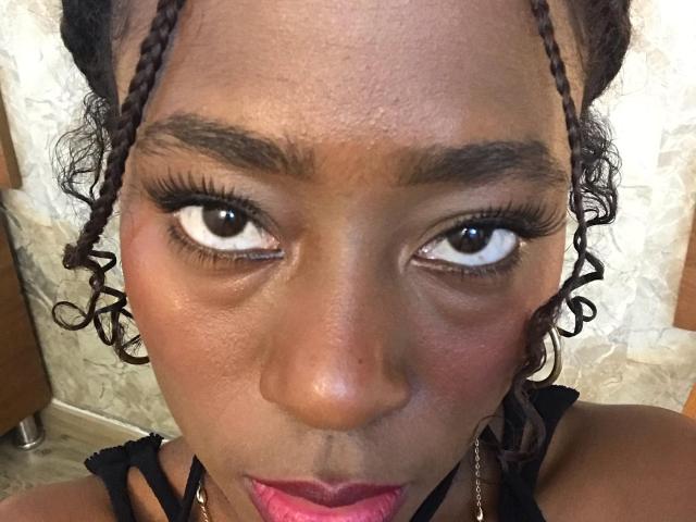 VioletGraice has dark brown skin, curly braided hair, thick eyebrows, long eyelashes, and pink lipstick. Her wide eyes and slightly puckered lips are centered in the image.