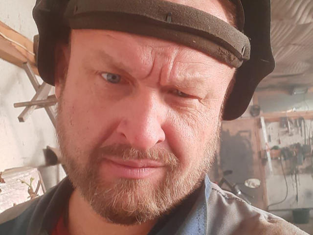 Grandor has a grumpy face, wearing a black metal helmet, with a thick beard and mustache, blue eyes, and a slightly furrowed brow, looking annoyed.