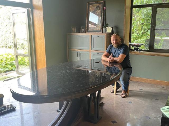 Grandor with a chubby ass, short blonde hair, and a blue shirt, standing by a curved black table, looking down with hands together.