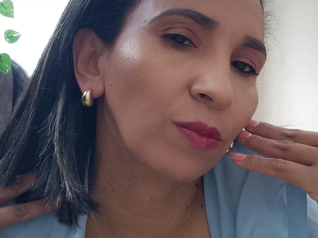 MrsVictoria's face, with medium brown skin, pink eyeshadow, red lipstick, and black shoulder-length hair, wearing gold earrings and a blue top, puckering her lips slightly.