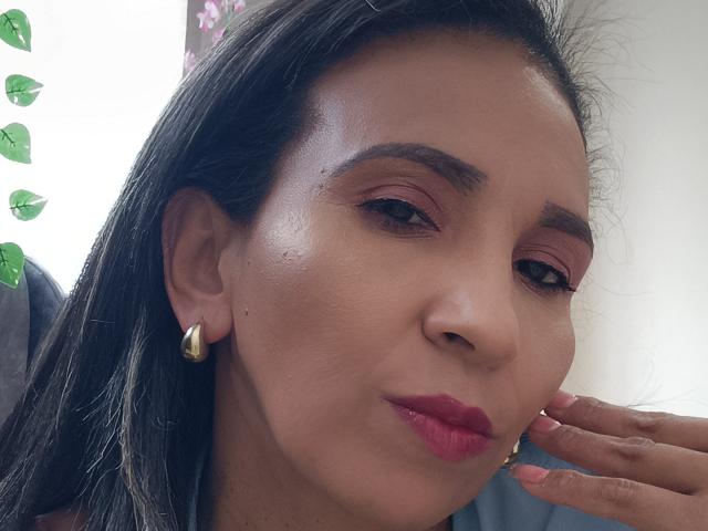 MrsVictoria has smooth brown skin, dark brown eyebrows, pink eyeshadow, red lipstick, small gold earrings, and is touching her cheek with her hand, showing a subtle smile.