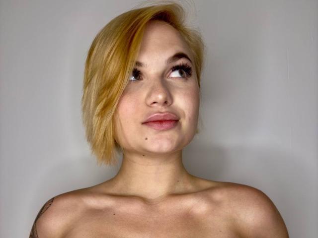 IvorySoul with short, blonde hair, looking up, smiling slightly, showing a small mole on her chin, and a tattoo on her right shoulder. She's topless with visible shoulders.