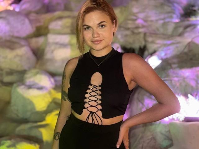 IvorySoul with tan skin, blonde hair, and tattoos, wearing a black cutout top showing her tits, lace-up front, and black pants, smiling at the camera.