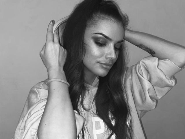 IngridI has long dark hair, wearing a loose shirt, making a peace sign with her right hand, her left hand in her hair, showing a tattoo on her forearm.