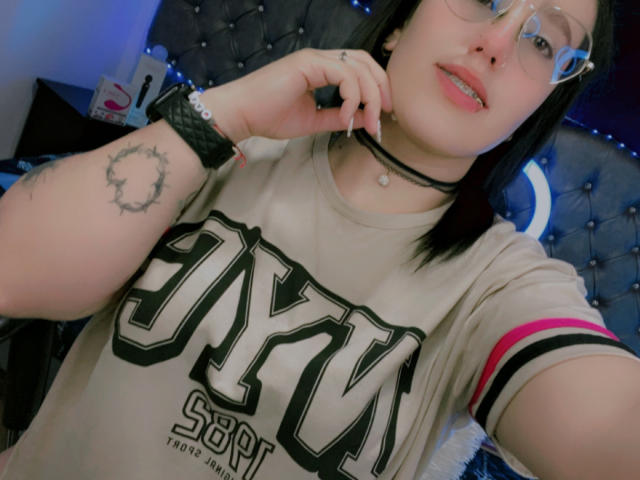 NatalieHarris with black hair, glasses, pink lip, choker, "SKY" tee, pink arm stripe, ring tattoo, black wristband, finger to mouth, smiling, blue light background.