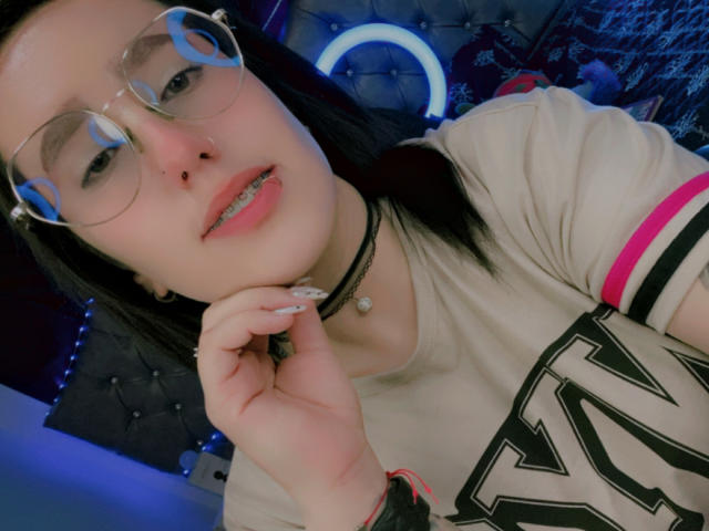 NatalieHarris with glasses, black hair, pink lip ring, and nose piercing, wearing a beige "K" shirt, black choker, and pink arm stripe, hand near mouth, looking at camera.