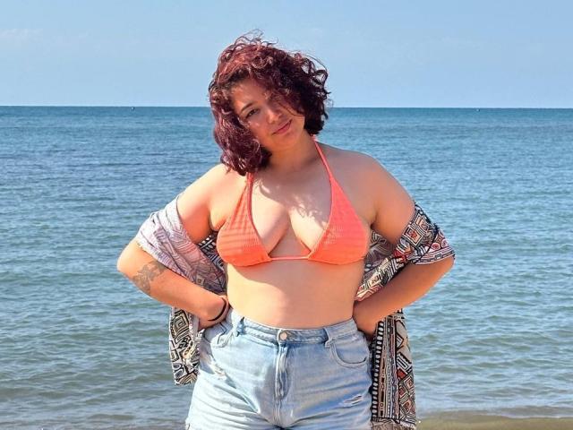 StarlitGaze has curly red hair, an orange bikini top showing her big tits, and light blue jeans with hands on her hips, standing by the ocean.